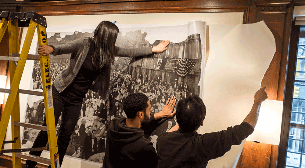 Photograph of staff on a ladder hanging a large photo in the Casa Italiana Salone gallery.